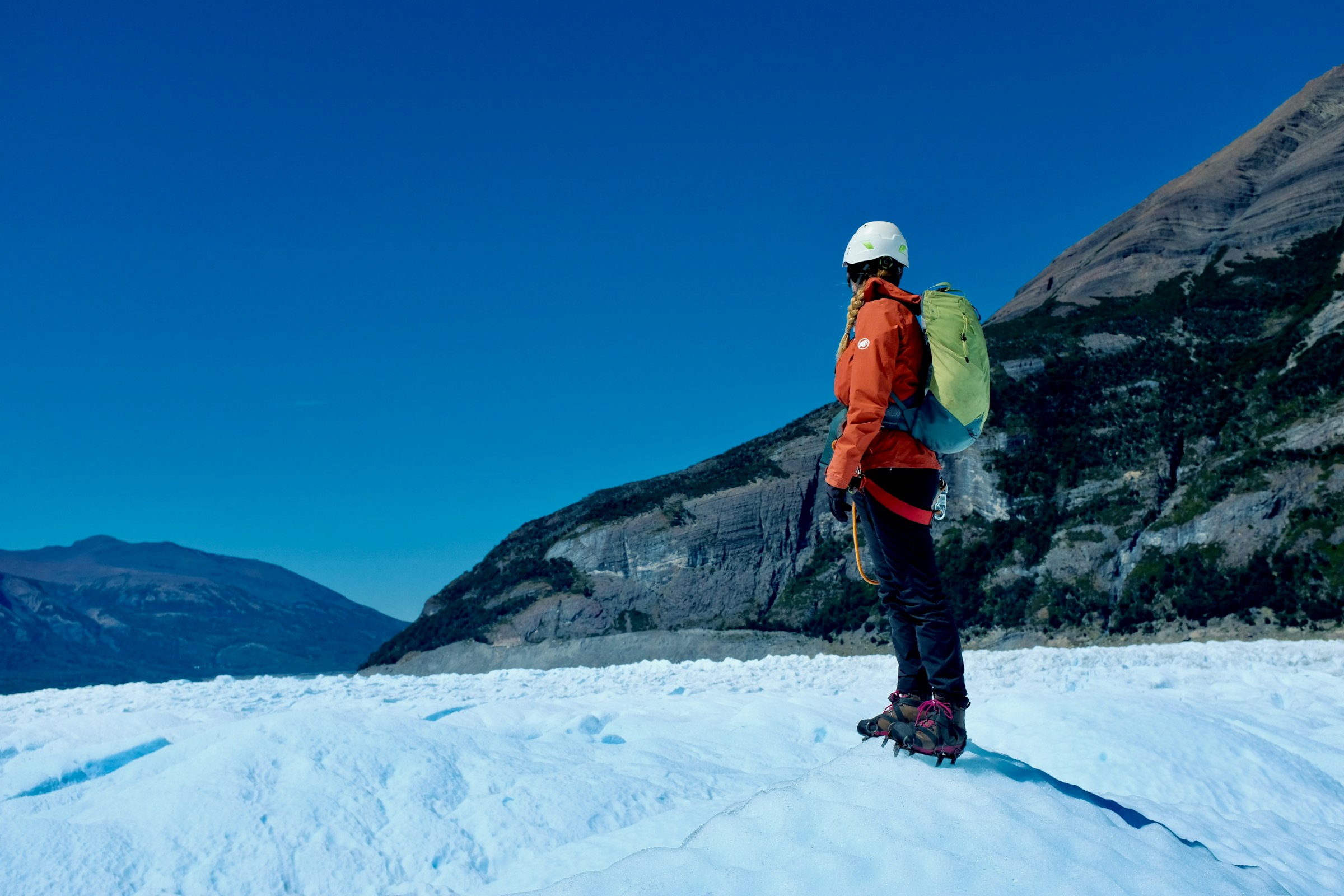 Climber in an orange jacket and white helmet standing on glacier ice beneath a dark mountain slope