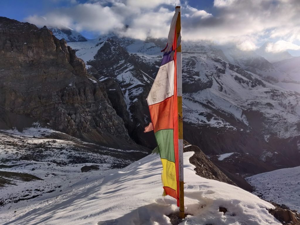 Tall pole of colourful prayer flags on a snowy ridge facing steep rock and snow slopes