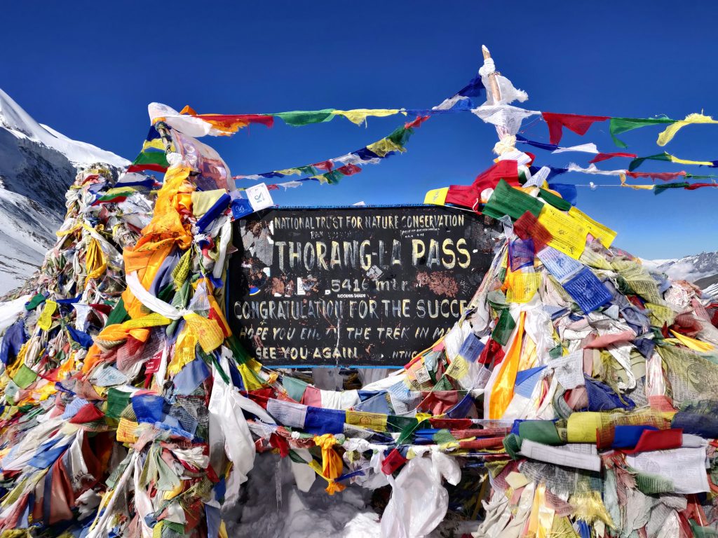 Thorong La Pass sign at 5,416 metres covered in colourful prayer flags under a blue sky