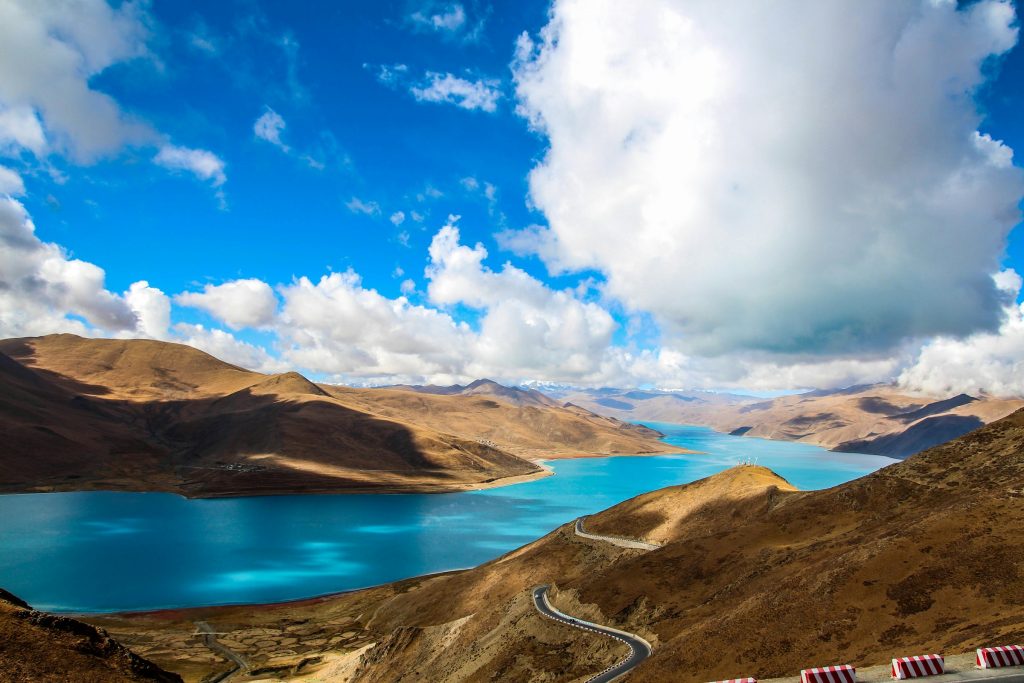 Turquoise Yamdrok Lake curving between brown hills with a winding road below a cloudy sky