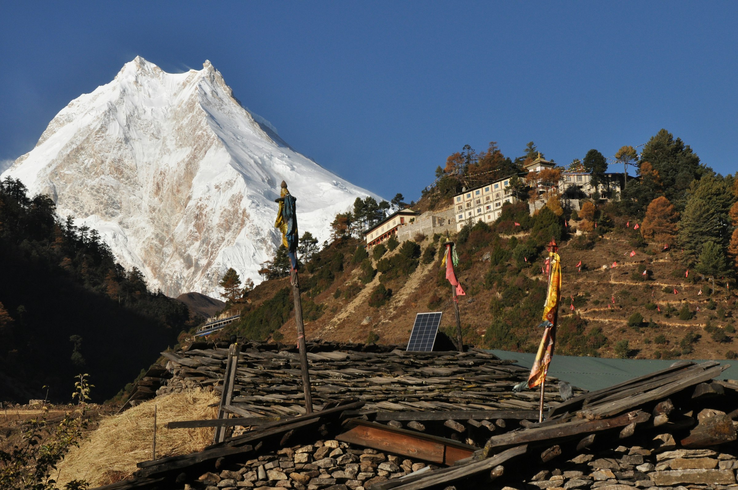 Manaslu's twin snowy summits above a hillside monastery and stone rooftops with prayer flags